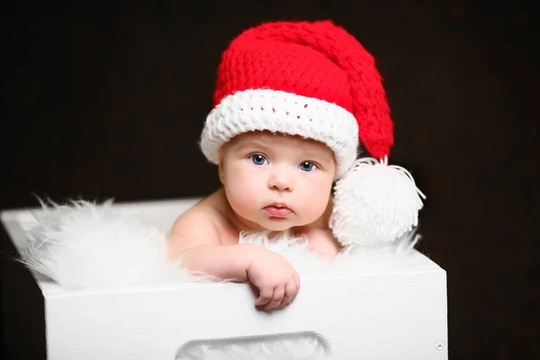 A karácsonyi baba ül egy fehér fa doboz, piros farkú kalapban — Stock Fotó