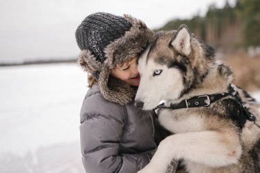 Kıvırcık saçlı küçük çocuk Sibirya köpeğine sarılıyor. 