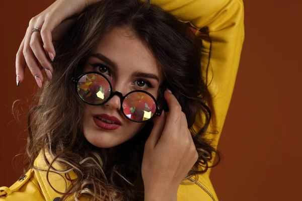 スタジオポートレートの美しい女の子で黄色のジャケットと眼鏡で色の眼鏡で茶色の背景 — ストック写真