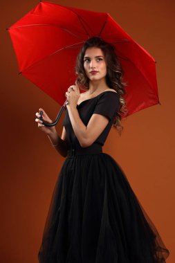 Açık kahverengi arka planda kırmızı şemsiyeli siyah elbiseli güzel bir kızın stüdyo portresi.