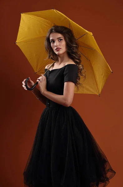 スタジオポートレートの美しい女の子で黒のドレスに黄色の傘の上にライトブラウンの背景 — ストック写真