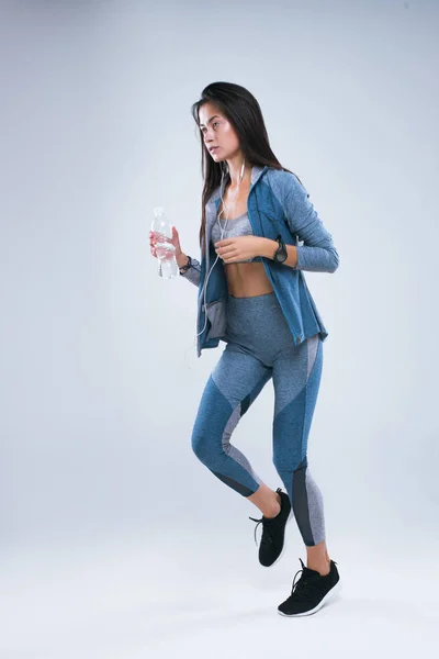 Hermosa chica del deporte correr con botella de agua en los auriculares y escuchar música. Longitud completa de la mujer de los deportes corriendo en el estudio sobre fondo gris — Foto de Stock