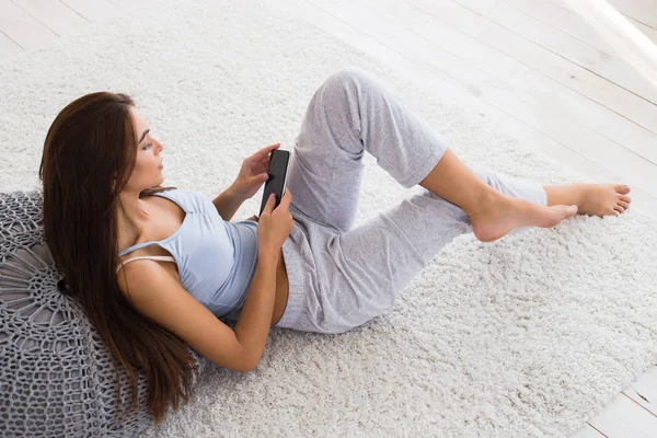 Красивая женщина расслабляется на полу и печатает сообщение. Женщина с помощью смартфона и укладки . — стоковое фото