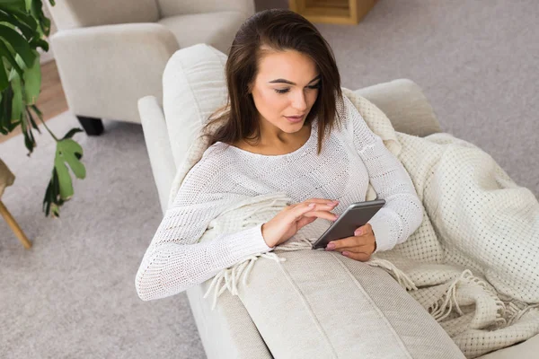Использование современных технологий дома. Красивая молодая женщина держит смартфон во время отдыха на диване дома — стоковое фото