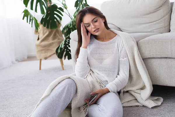 Baş ağrısı. Kanepede evde oturan genç bir kadın elini kafasına koyar.