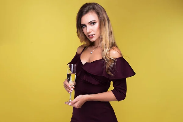 Jovem loira sexy segurando uma taça de champanhe, olhando para a câmera e sorrindo. Mulher atraente em vestido sexy no estúdio no fundo amarelo — Fotografia de Stock