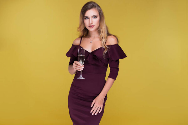 Sexy blond young woman holding a glass of champagne, looking at the camera and smile.  Attractive woman in sexy dress in studio on yellow background