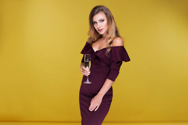 Sexy blond young woman holding a glass of champagne, looking at the camera and smile.  Attractive woman in sexy dress in studio on yellow background