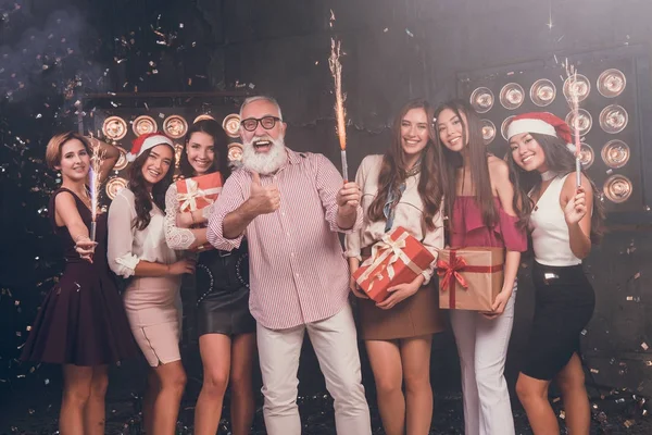 Santa give gifts for girls. Modern bearded man showing OK while carrying Christmas bengal lights with beautiful girls in club with gold confetti celebrate New Year and Christmas party 2018 — Stock Photo, Image