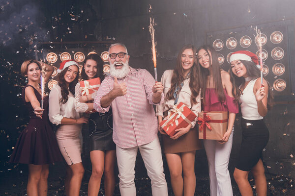  Santa give gifts for girls. Modern bearded man showing OK while carrying Christmas bengal lights with beautiful girls in club with gold confetti celebrate New Year and Christmas party 2018