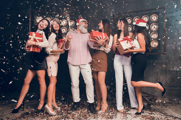 Santa give gifts for girls.  Modern bearded man in Santa's hat carrying Christmas bengal lights with beautiful girls in club with gold confetti celebrate New Year and Christmas party 2018