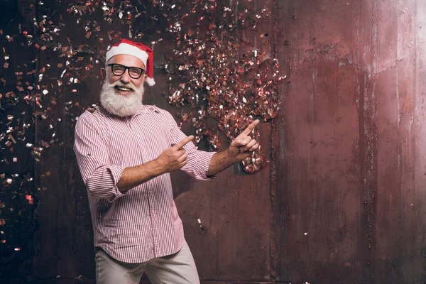 Happy bearded and modern man in Santa's hat and in fashionable glasses shows his fingers to the side and happy smile in gold confetti. Happy New Year 2018 and modern party with fashion Santa — Stock Photo, Image