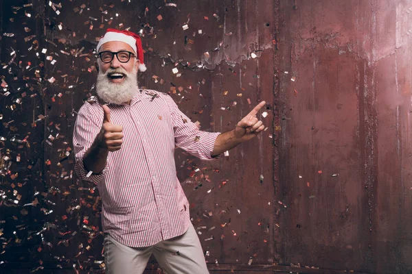 Feliz hombre barbudo y moderno en el sombrero de Santa y en gafas de moda muestra sus dedos a un lado y feliz sonrisa en confeti de oro. Feliz Año Nuevo 2018 y fiesta moderna con la moda de Santa — Foto de Stock