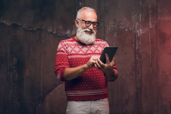 Un hombre mayor feliz moderno usando un teléfono inteligente en un suéter de Navidad. Santa Claus desea Feliz Navidad —  Fotos de Stock