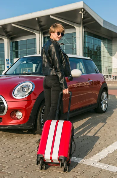 Όμορφο κορίτσι κοιτάζοντας την κάμερα μένοντας κοντά στο αυτοκίνητο. Κορίτσι πρόκειται για διακοπές με την κόκκινη βαλίτσα. Καλοκαιρινή διάθεση, νέες συγκινήσεις από μελλοντικές διακοπές. — Φωτογραφία Αρχείου
