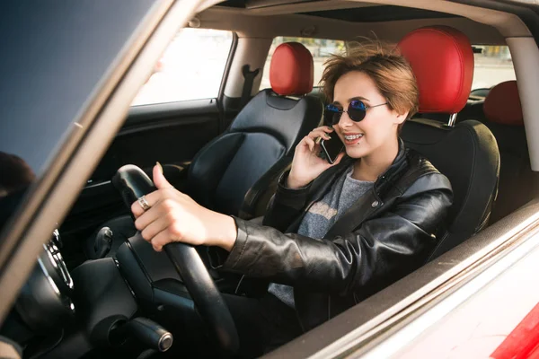 Hermosa mujer de negocios en chaqueta de cuero está hablando en el teléfono móvil y sonriendo mientras conduce en un coche. Feliz día ocupado en una gran ciudad . — Foto de Stock