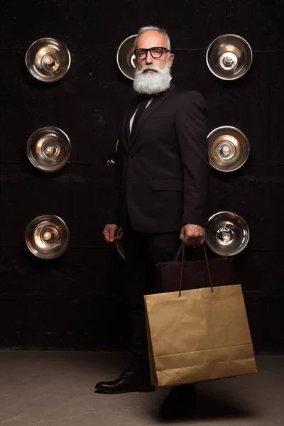 Ernsthafter Geschäftsmann nach dem Einkauf, der zwei große Päckchen mit neuer Mode bei sich trägt. Bärtiger Mann im dunklen Anzug posiert mit Einkäufen — Stockfoto