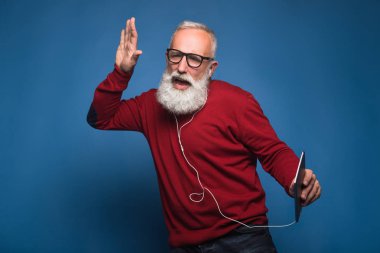 Yeni bir şarkı beklentisi. Tabletten yeni bir müzik dinlerken mutlu modern sakallı adam. Adam kulaklık ile mavi arka plan üzerinde izole müzik dinlemek. Mutlulukla cool müzik dansları kulaklıklar üzerinde.