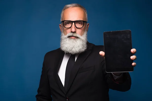 El hombre barbudo mayor concentrado en ropa formal con emociones serias sostiene la tableta digital y muestra espacio de copia en la pantalla mientras está de pie sobre fondo azul. Lugar vacío de la pantalla de la tableta —  Fotos de Stock