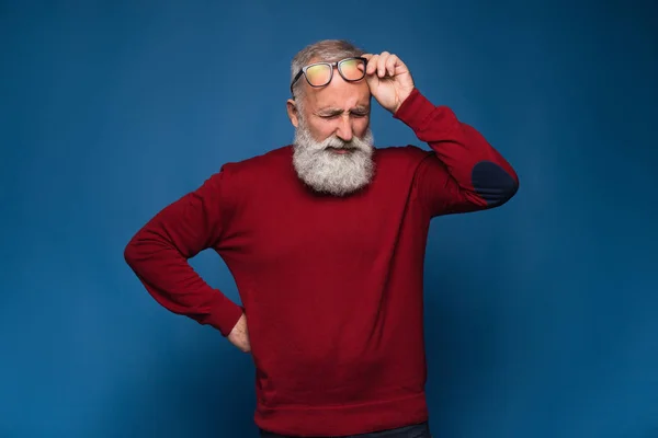 Headache. Old bearded man in red sweeter and in black glasses show headache. Pain emotions isolate on blue background. Stress after hard working office day — Stock Photo, Image