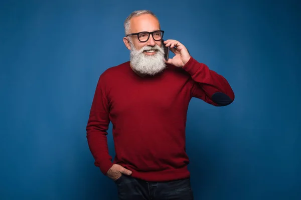 Usando smartphone aislado sobre fondo azul. Feliz conwersation a los amigos por el hombre barbudo en rojo más dulce y gafas de moda. Hombre hablando por teléfono y sonriendo mientras mira hacia otro lado —  Fotos de Stock