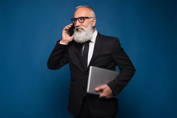 Ocupado barbudo senior y hombre de negocios que se queda con portátil y teléfono móvil aislado sobre un fondo azul . —  Fotos de Stock