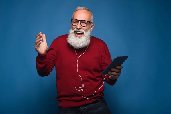 Esperanza de una nueva canción. Feliz hombre barbudo moderno escuchando una nueva música de la tableta. El hombre con auriculares escucha música aislada sobre fondo azul. Felizmente baila de música fresca en los auriculares . —  Fotos de Stock