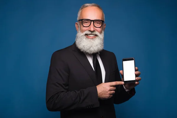 Hombre barbudo feliz en ropa formal sostiene el teléfono móvil y muestra espacio de copia en la pantalla mientras está de pie sobre fondo azul. Lugar vacío de la pantalla del teléfono . —  Fotos de Stock