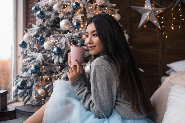 С Новым годом! красивая азиатская девушка сидит рядом с елкой и пьет кофе . — стоковое фото