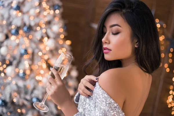 Mujer sexy con una copa de champán. Hermosa chica en un vestido sexy. ¡Brillante y alegre Navidad! Chica asiática en vestido de oropel plateado. Deseos más cálidos para Navidad ! —  Fotos de Stock