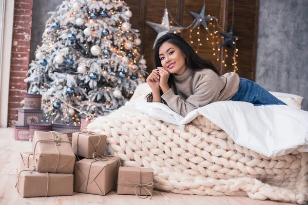 Gelukkig kerstochtend in een huis. Koude winter. Mooie Aziatische meisje in een zoeter en jeans opleggen de wit beddengoed slecht. Happy New Year's ochtend. Warmste wensen voor Kerstmis! — Stockfoto