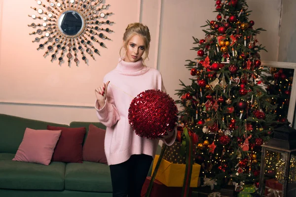 Beutiful blond and attractive woman with a red Christmas boll in her hands smile and look at the camera. Celebration New Year with fashion Christmas toys and happy mood.