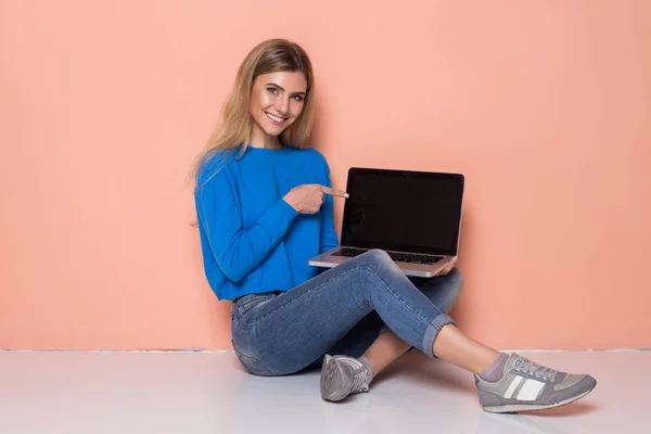 Улыбающаяся случайная женщина показывает чистый экран ноутбука на фоне оранжевого цвета. Смотреть в камеру — стоковое фото