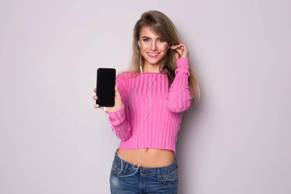 Femme blonde souriante en chemisier rose montrant écran de smartphone vierge et écoutant de la musique dans des écouteurs isolés sur un fond gris — Photo