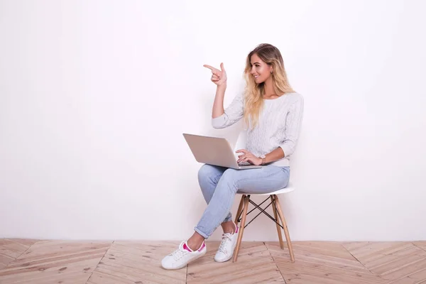 Молодая женщина сидит на стуле и работает над ноутбуком. И показывает руку в сторону — стоковое фото