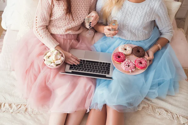 Duas meninas estão sentadas na cama enorme. Estão a ver filmes no portátil. Comer donuts e beber champanhe . — Fotografia de Stock