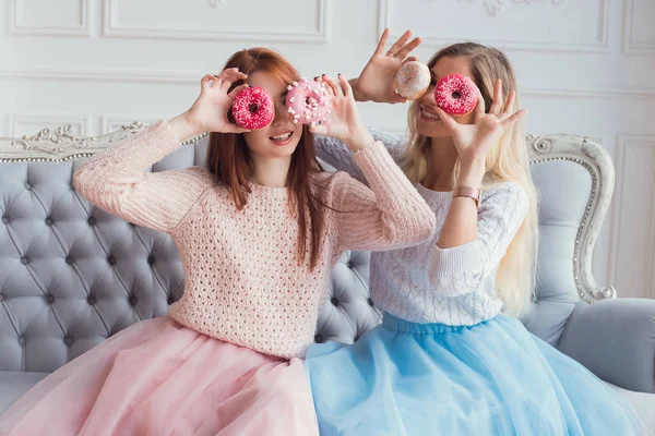 Duas meninas estão sentadas no sofá se divertindo. Eles estão segurando donuts na frente de seus olhos. Celebração da festa . — Fotografia de Stock