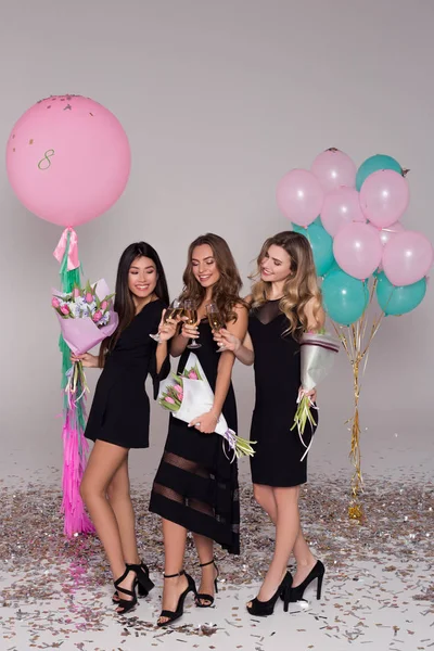 Três meninas bonitas em vestidos pretos estão segurando buquês de tulipa. As senhoras estão a beber champanhe. Balões coloridos. Confetti. . — Fotografia de Stock