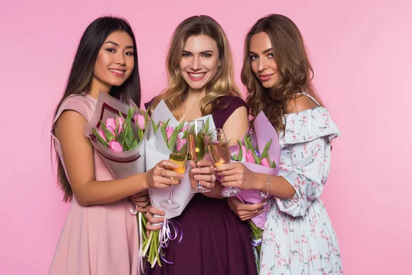 Drei schöne Mädchen in bunten Kleidern halten Tulpensträuße in der Hand. Damen trinken Champagner. rosa Hintergrund. — Stockfoto