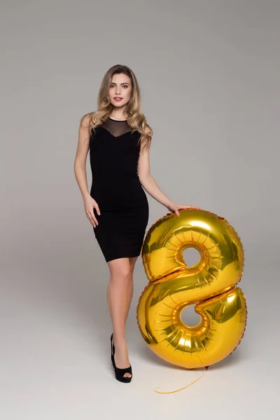 Sexy, hermosa chica en vestido negro está sosteniendo gran globo de oro en forma de 8. Fondo gris. Feliz día de la mujer —  Fotos de Stock