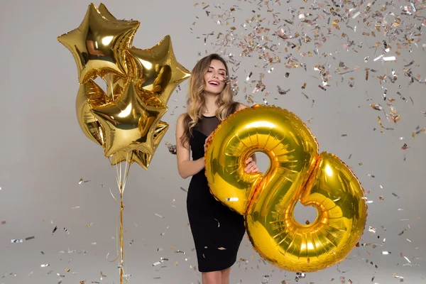 Menina bonita feliz em vestido preto está segurando grande balão dourado. Balões em forma de estrela. Confetti. Dia da mulher feliz — Fotografia de Stock