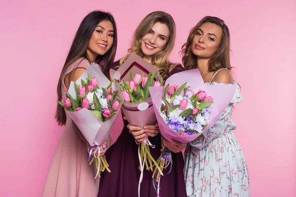 Drei schöne Mädchen in bunten Kleidern halten Tulpensträuße in der Hand. rosa Hintergrund. — Stockfoto