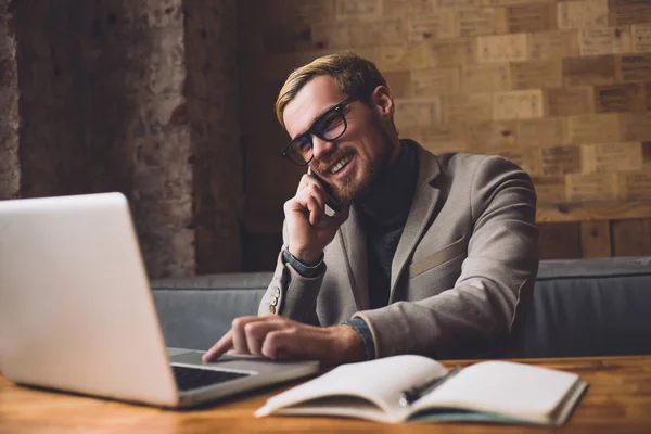 Hombre de negocios sonriente sentado junto a la mesa en la cafetería con computadora portátil y teléfono inteligente —  Fotos de Stock