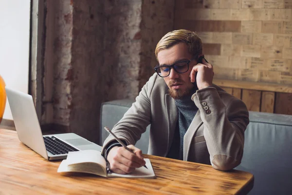 Joven hombre de negocios está tomando notas mientras habla por teléfono y trabajando en el portátil —  Fotos de Stock