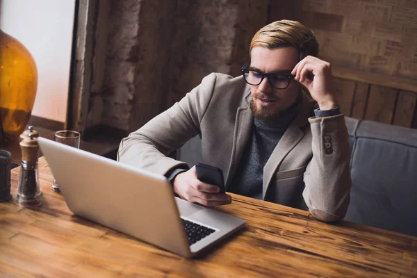Hombre de negocios guapo sentado junto a la mesa en la cafetería con ordenador portátil y teléfono inteligente —  Fotos de Stock