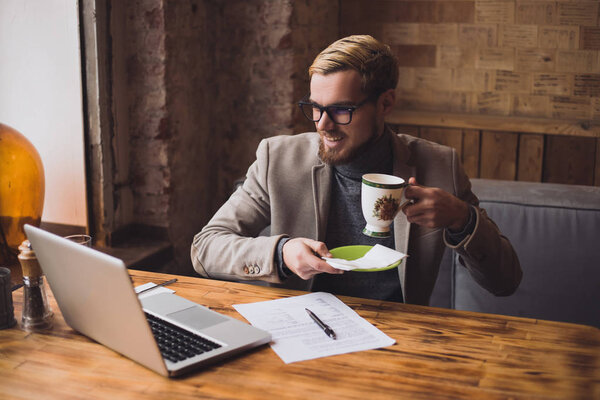 Young man working at the laptop and drinking a coffee during a business video conversation 