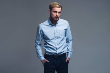 Ciddi ve yakışıklı. Kollarını cepler gri arka plan önünde tutan genç iş adamı.