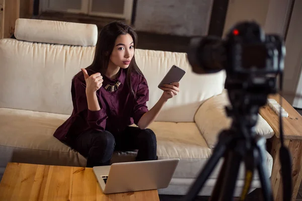 Красивая молодая девочка-блогер сидит на диване со своим смартфоном и ноутбуком и ведет блог, используя камеру с треногой . — стоковое фото