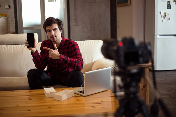 Joven hombre barbudo inteligente haciendo un video para su propio blog y la publicidad del teléfono inteligente en el sofá de cuero blanco en casa . —  Fotos de Stock