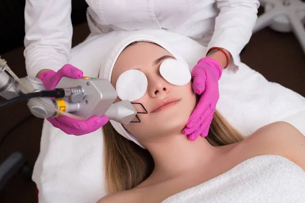 Take care of yourself! Close-up top-view portrait of a young girl with a towel on her head lying on a table with closed eyes getting a laser skin treatment in healthy beauty spa salon.